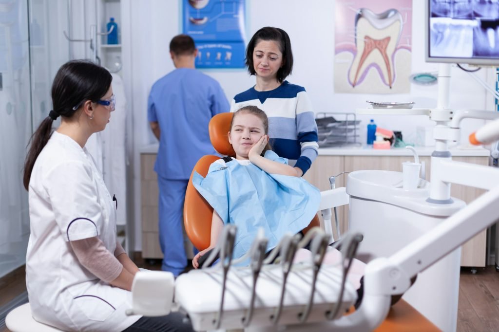 Little girl with painful expression showing dentist where her tooth hurts